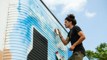 Portrait of Jeremy Flores standing with spray paint in hand on ladder painting clouds on structure.