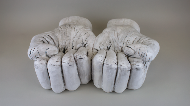 A photo of a plaster sculpture of two hands in upward facing fists sitting on a gray backdrop.