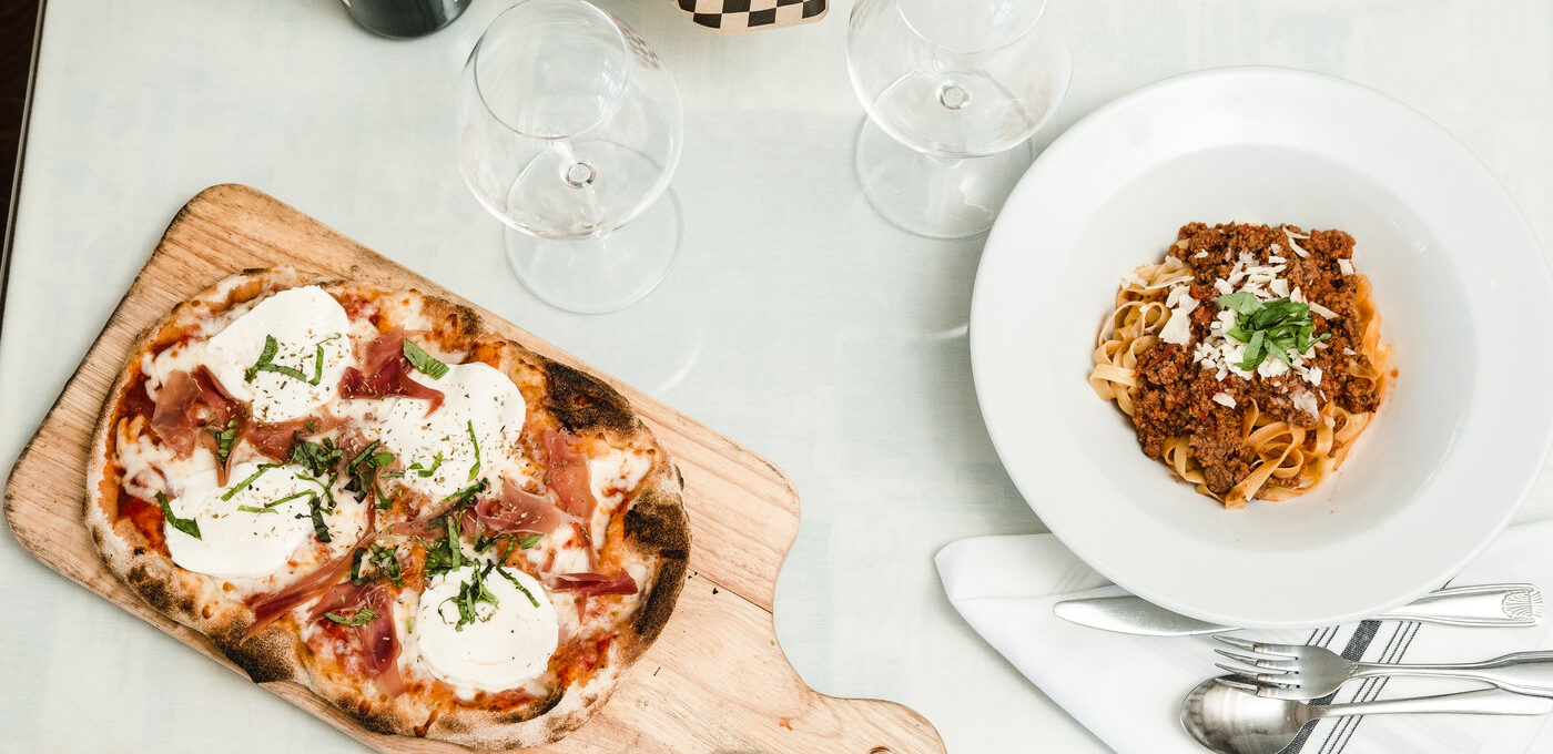 A wine bottle and two glasses next to a pizza and pasta. Photo By Rachel Benavides