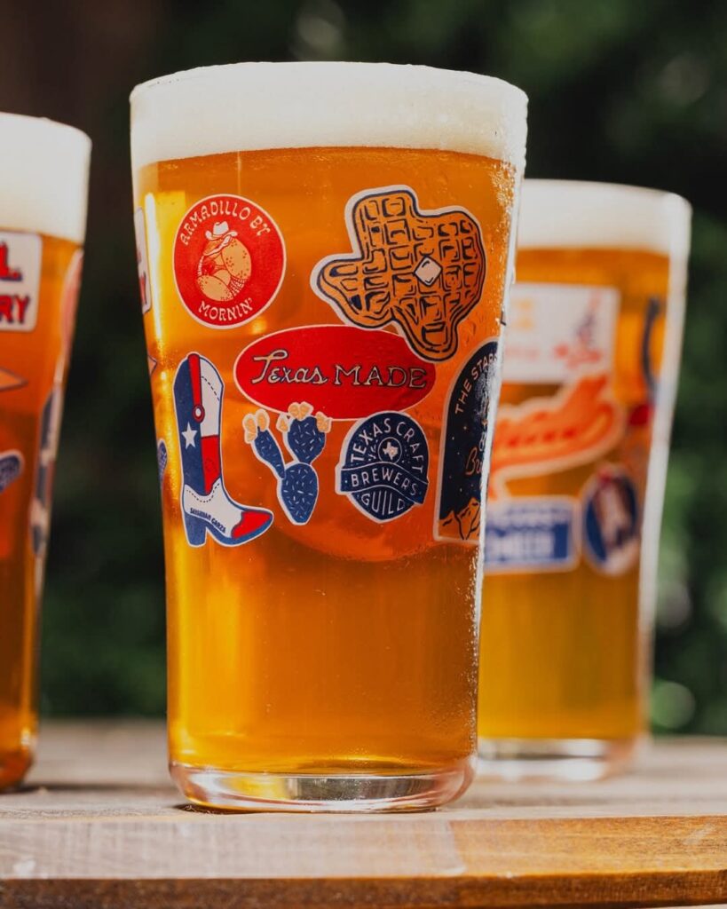 An image of a glass pint filled with beer and covered in faux Texas-theme stickers, designed by Savannah Garza for the 2025 Small Business Saturday limited edition pint glass.