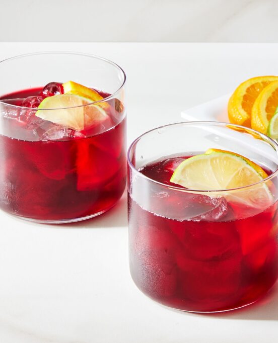 Two glasses of hibiscus tea with slices of lemon floating on top sit on a white countertop.