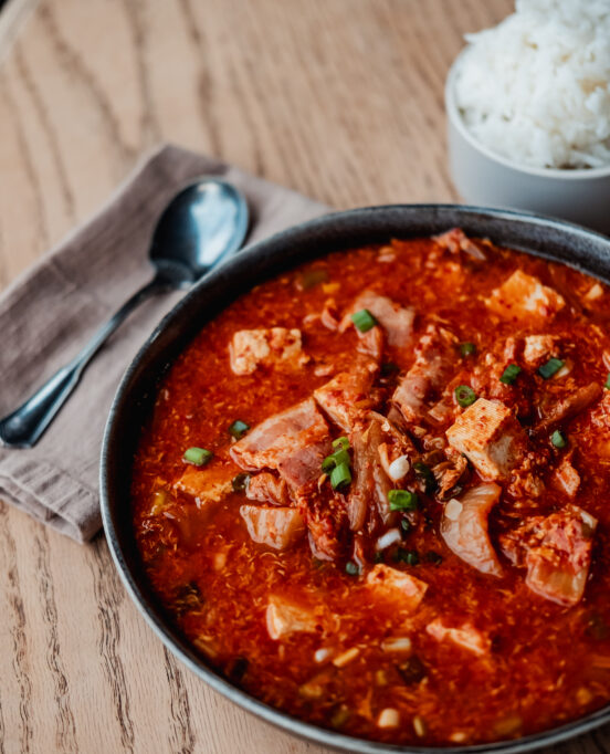 A close-up on ZCounter's kimchi soup, served inside a black bowl next to a bowl of rice and a spoon and napkin.