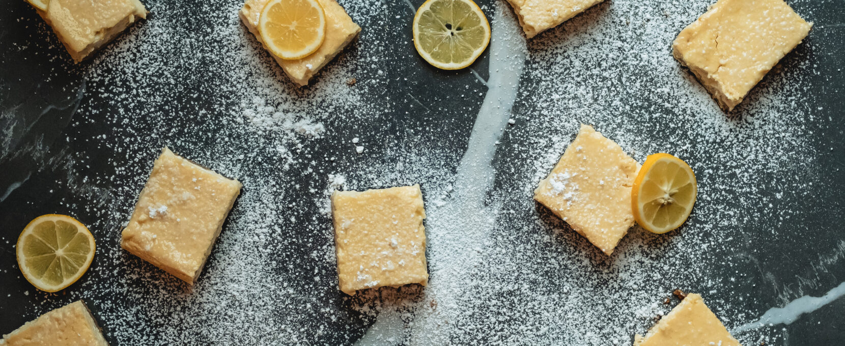 An overhead view of lemon bars and lemon slices sit scattered across a marble counter top sprinkled with powdered sugar.