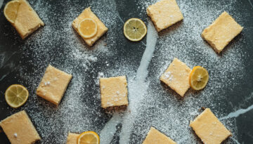 An overhead view of lemon bars and lemon slices sit scattered across a marble counter top sprinkled with powdered sugar.
