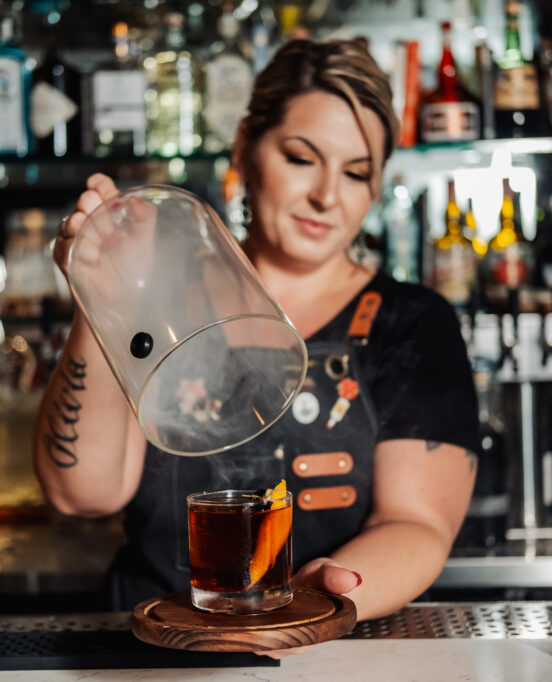 A female bartender unveils a dark craft cocktail in a small air of smoke inside Fork & Vines.