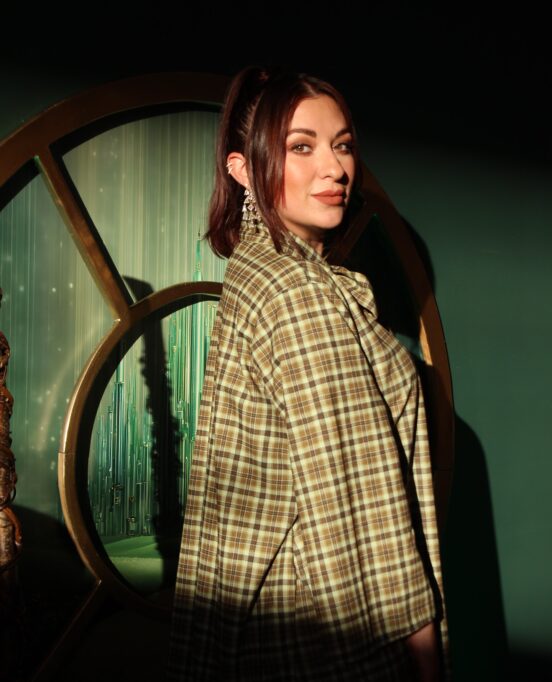 Clarissa Serna poses in front of a green background with a plaid outfit on.