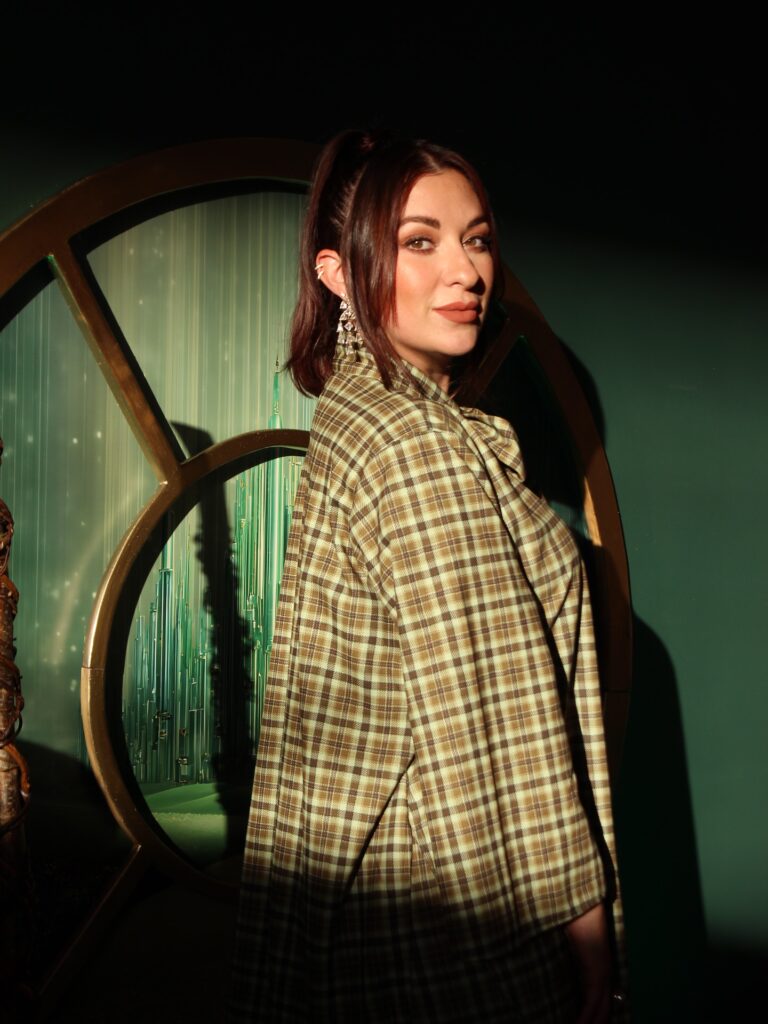 Clarissa Serna poses in front of a green background with a plaid outfit on. 