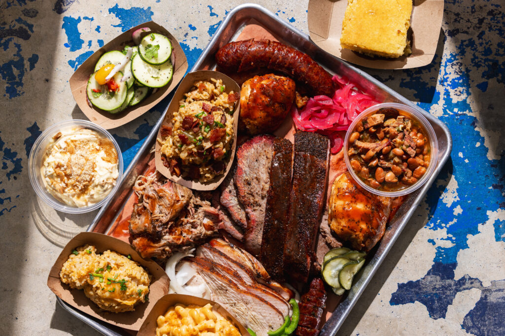 The photo features a barbeque spread of ribs, sausage, corn and other various sides.