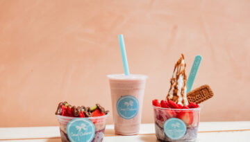 Three menu items from Coco Cabana sit on a white table against a soft pink background. To the left is an acai bowl, in the center is a smoothie and on the right is a soft-serve.