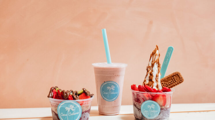 Three menu items from Coco Cabana sit on a white table against a soft pink background. To the left is an acai bowl, in the center is a smoothie and on the right is a soft-serve.