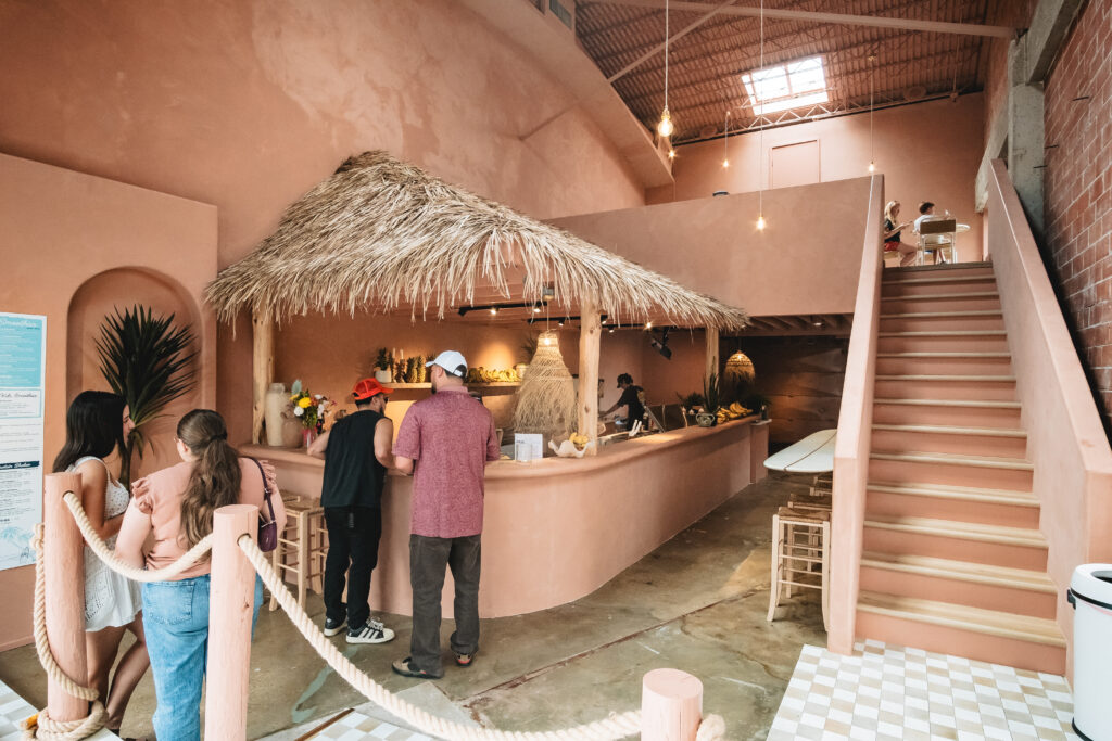 The photo features the inside of the Coco Cabana downtown location. In the center of the photo, the bar can be seen surrounded by people as they pick up their orders. To the right there is a staircase leading to the second level of the building. The walls are a soft, warm pink color.