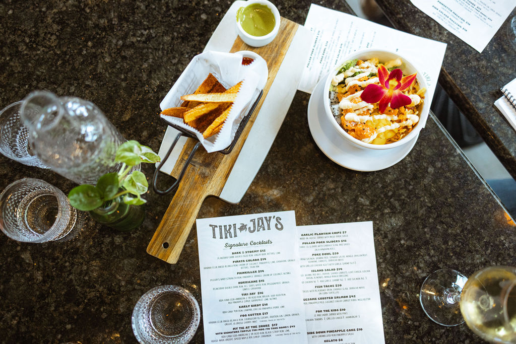 The photo is taken against a brown marble table at birds eye view. To the left, drink glasses sit, accompanied by a small plant. In the center, there is a wooden slab that holds a container of french fries and a sauce, there is a poke bowl in white ceramic with a pink flower on top and a menu for Tiki jays.