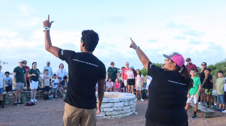 Camp Aranzazu counselors face a group of campers and hold their hands up to the sky. The back of their shirts are in focus and say "A world built for belonging."