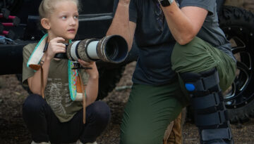 Sharp Shooter Foto Photographer shows young girl how to hold camera. The two are crouched in front of a vehicle, ready to capture a photo of nature.