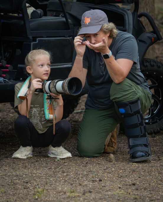 Sharp Shooter Foto Photographer shows young girl how to hold camera. The two are crouched in front of a vehicle, ready to capture a photo of nature.