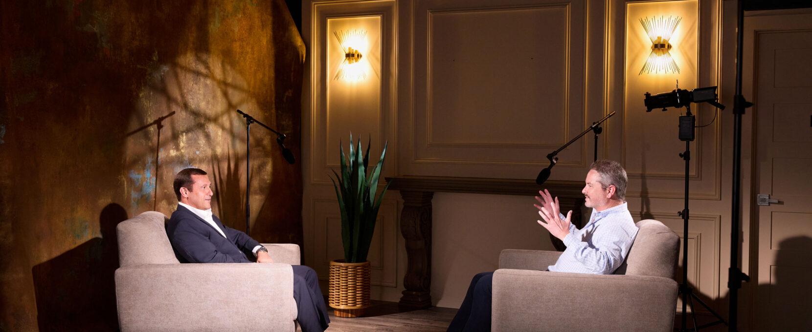 Nathan Swinney (left) and Stephen Levy (right) face each other in grey armchairs as they talk about what makes a construction project successful. Cameras and microphones surround them. Behind Nathan Swinney sits a bronze drape background.