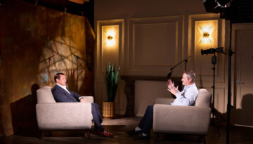 Nathan Swinney (left) and Stephen Levy (right) face each other in grey armchairs as they talk about what makes a construction project successful. Cameras and microphones surround them. Behind Nathan Swinney sits a bronze drape background.
