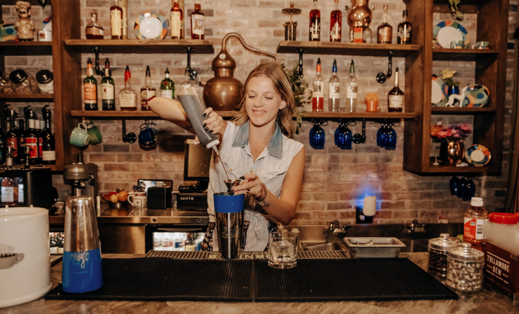 A female bartender mixes a drink behind a bar. Behind her are shelves lined with alcohol and beverage-making tools. 