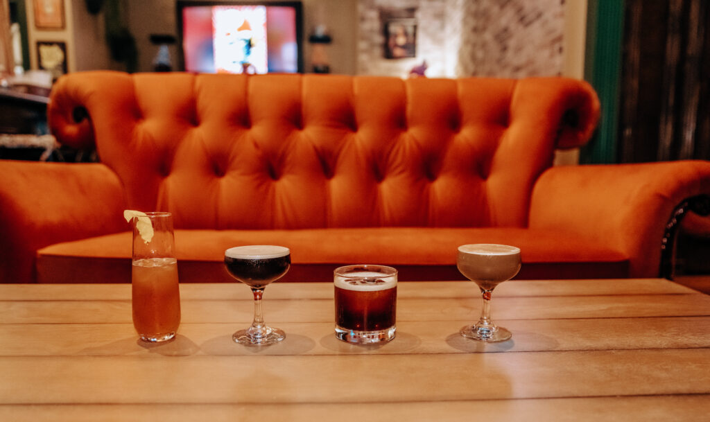 The photo features four specialty drinks sitting on a table. Behind the table is an orange couch. 