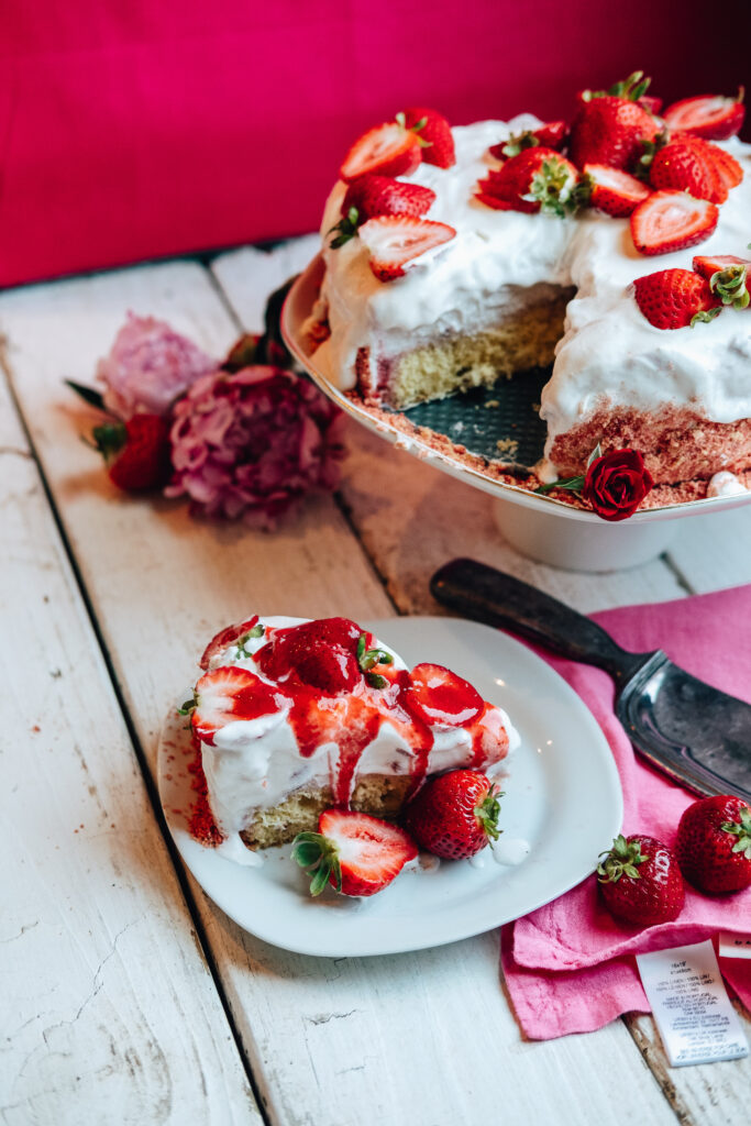 The Strawberry Ice Cream Shortcake sits on a cake display with a slice cut out, sitting on a separate plate. The cake is dressed with strawberries on top, and the slice has strawberry syrup drizzled on top.