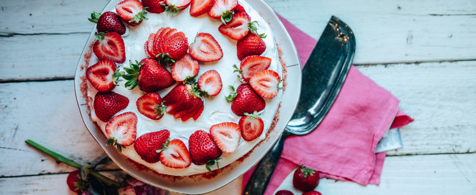 The Strawberry Ice Cream Shortcake sits on a cake stand. The photo in birds eye view shows the cake dressed in strawberries, surrounded by flowers, strawberries, a pink napkin and a cake cutter.