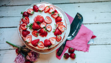 The Strawberry Ice Cream Shortcake sits on a cake stand. The photo in birds eye view shows the cake dressed in strawberries, surrounded by flowers, strawberries, a pink napkin and a cake cutter.