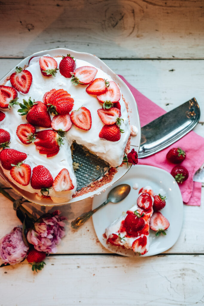 The photo is taken through birds eye view. It features the cake with a slice missing. The missing slice is sitting next to the cake on a plate and has strawberry syrup drizzled on the top and is joined by a spoon hanging off the side of the plate. Under the cake stand, the cake cutter can be seen laying on top of a pink napkin next to strawberries and flowers.