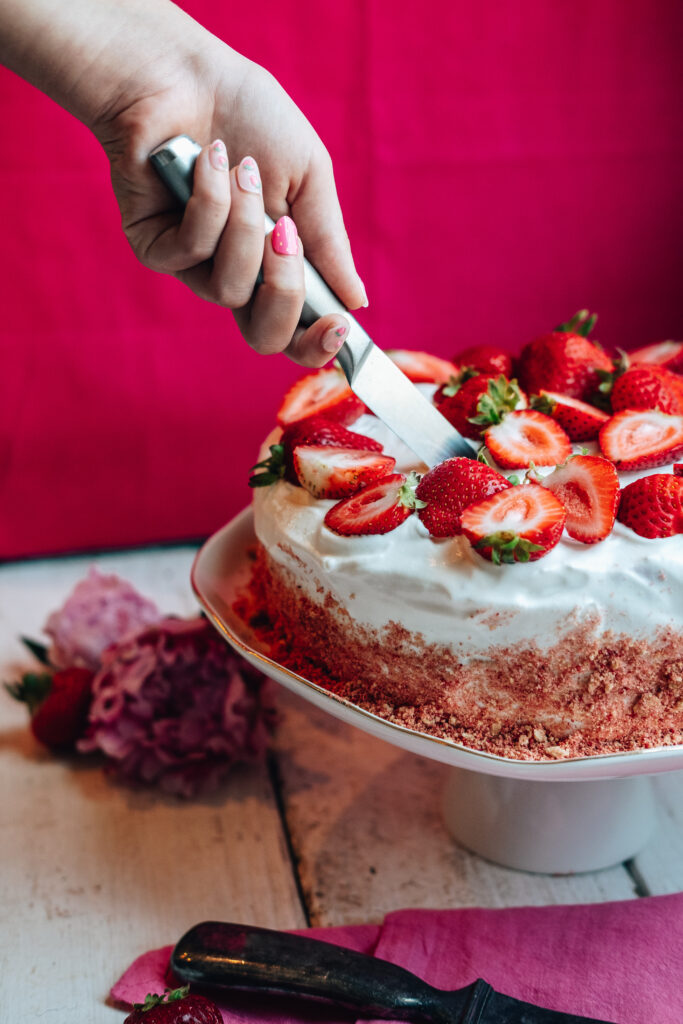 A hand hold's a knife as it cuts into the cake. The cake sits atop a cake stand on a white table. The background is a vibrant magenta pink.