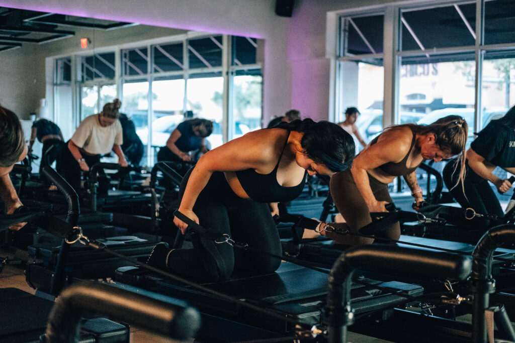 A woman is featured doing a Pilates exercise with the Xformer machine. She is in the Pilates studio, surrounded by other women doing the same exercise. Behind her, there is a mirror that sits against the purple walls. On the wall to the right there are windows.