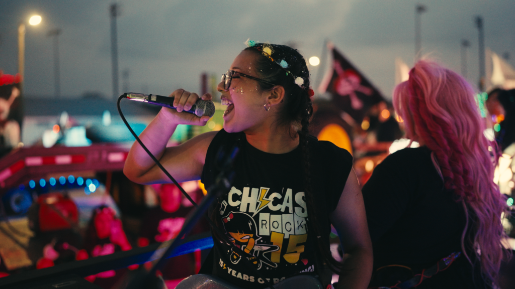 A young girl sings into a microphone on stage. wearing a Chicas Rock shirt.