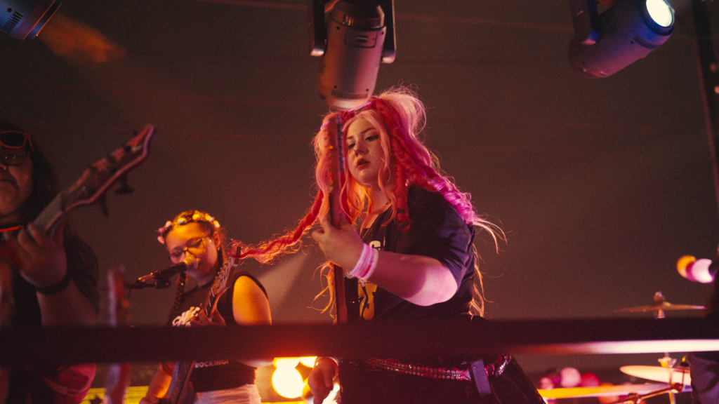 A young girl with pink hair plays the guitar on stage.
