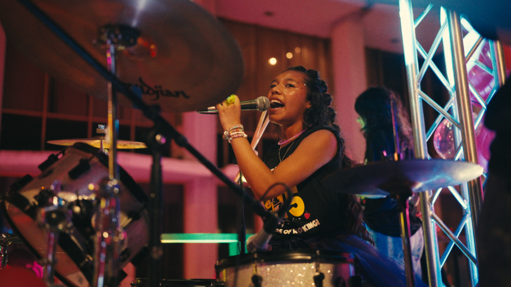 A young girl sits at a drum set, singing into a microphone.